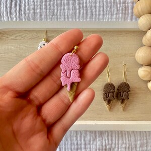 May include: A pair of gold hoop earrings with a pink and brown ice cream cone charm. The ice cream cone charm is made of polymer clay and has a textured surface. The earrings are a fun and whimsical accessory for any occasion.