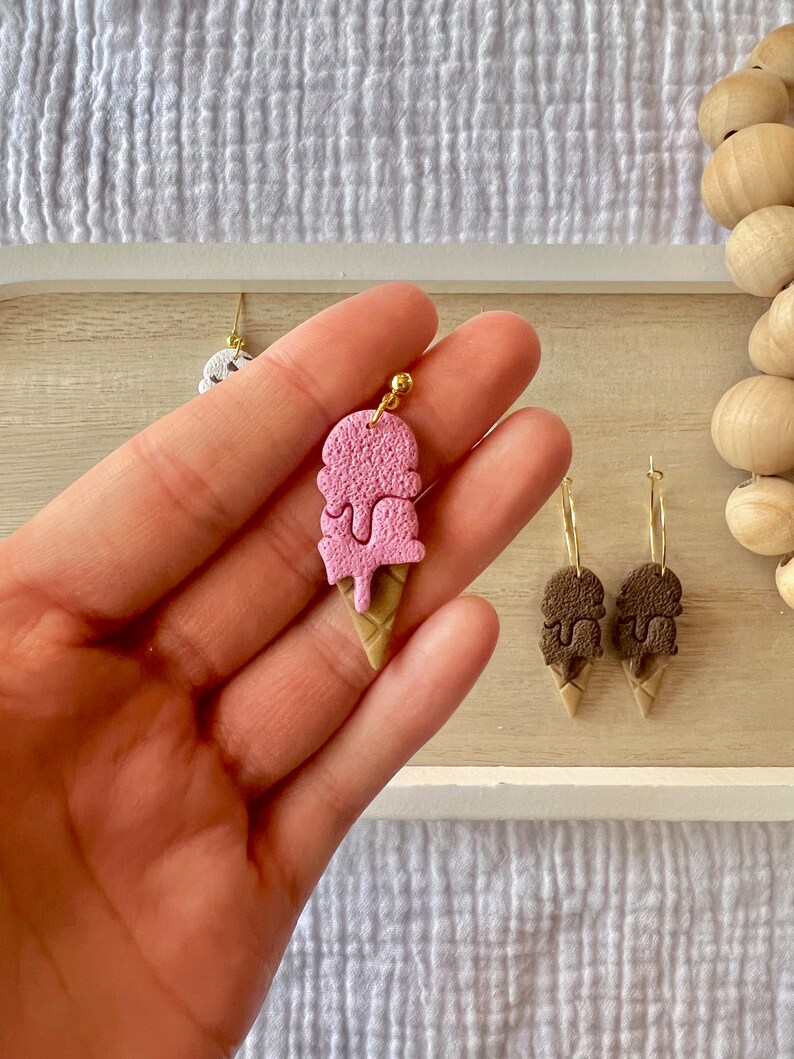 May include: A pair of gold hoop earrings with a pink and brown ice cream cone charm. The ice cream cone charm is made of polymer clay and has a textured surface. The earrings are a fun and whimsical accessory for any occasion.