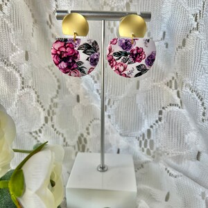 May include: A pair of gold and white floral earrings with a pink and purple floral pattern. The earrings are on a white stand.