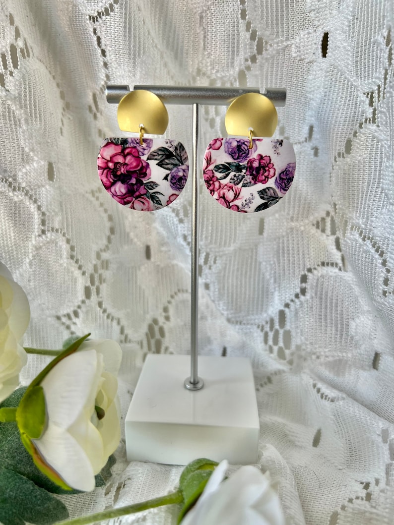 May include: A pair of gold and white floral earrings with a pink and purple floral pattern. The earrings are on a white stand.