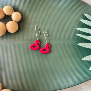 Peut inclure: Une paire de boucles d'oreilles flamant rose vif avec des crochets dorés. Les boucles d'oreilles sont présentées sur un plateau vert en forme de feuille, avec des perles en bois en arrière-plan. Un accessoire amusant sur le thème tropical.