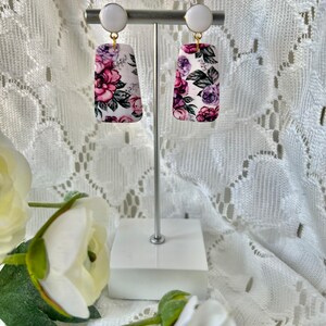 May include: A pair of floral print earrings with white and gold accents. The earrings are rectangular in shape and feature a pink, purple, and white floral pattern.