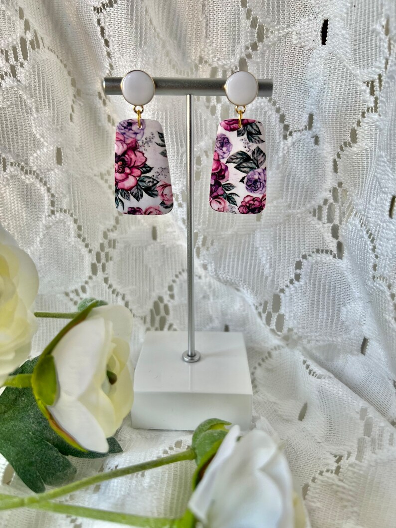 May include: A pair of floral print earrings with white and gold accents. The earrings are rectangular in shape and feature a pink, purple, and white floral pattern.