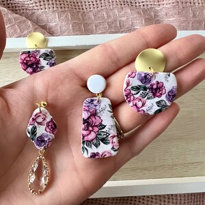May include: Three pairs of floral print earrings with gold accents. The earrings are made of polymer clay and feature a white background with pink and purple flowers. One pair has a teardrop-shaped crystal dangle.