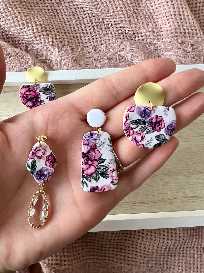 May include: Three pairs of floral print earrings with gold accents. The earrings are made of polymer clay and feature a white background with pink and purple flowers. One pair has a teardrop-shaped crystal dangle.