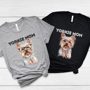 May include: Two t-shirts with the text "Yorkie Mom" printed on them. One shirt is grey and the other is black. Both shirts feature a cartoon image of a Yorkshire Terrier wearing glasses and a bow tie.