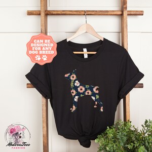 Puede incluir: Camiseta negra con una silueta de Doberman con estampado floral. El texto "Se puede dise&ntilde;ar para cualquier raza de perro" est&aacute; en un c&iacute;rculo rosa con huellas de patas.