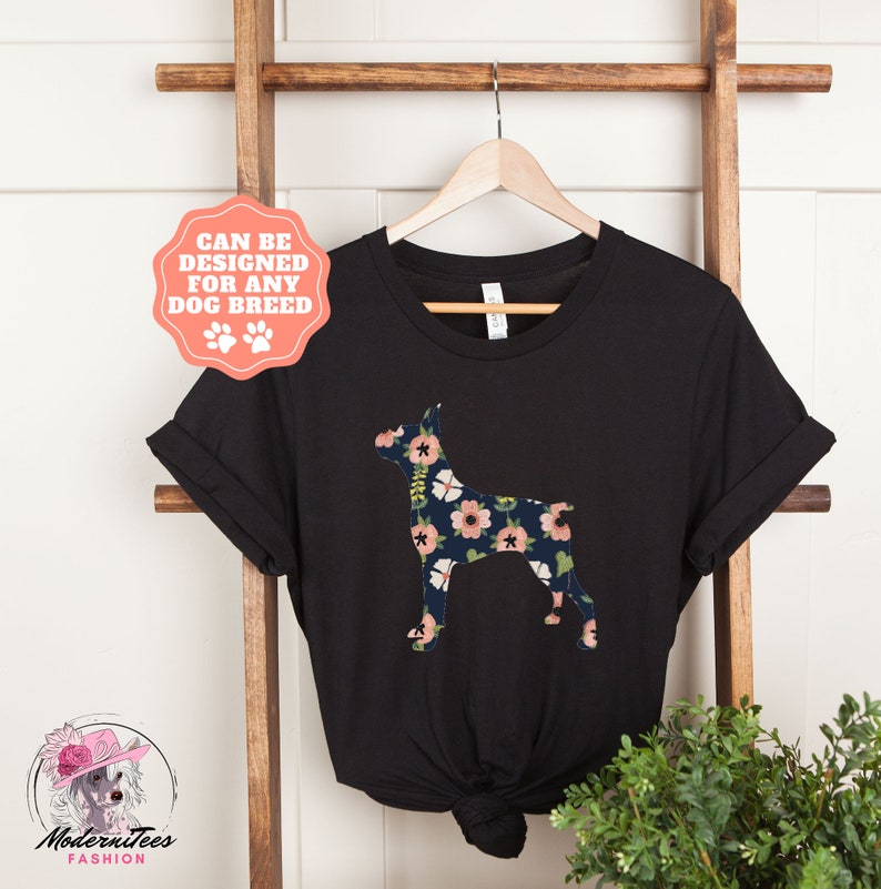 Puede incluir: Camiseta negra con una silueta de Doberman con estampado floral. El texto "Se puede dise&ntilde;ar para cualquier raza de perro" est&aacute; en un c&iacute;rculo rosa con huellas de patas.