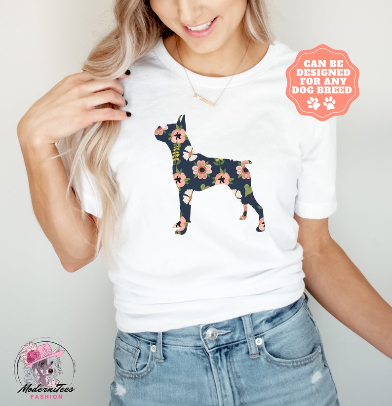 Puede incluir: Una camiseta blanca con un estampado floral de una silueta de Doberman Pinscher. El texto "Se puede dise&ntilde;ar para cualquier raza de perro" est&aacute; en una pegatina rosa con forma de huella de pata.