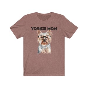 May include: A heather brown t-shirt with the text "Yorkie Mom" printed in black. A watercolor illustration of a Yorkshire Terrier wearing glasses is centered on the shirt.