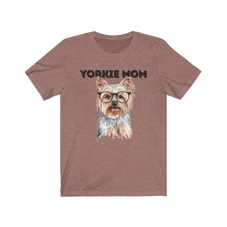 May include: A heather brown t-shirt with the text "Yorkie Mom" printed in black. A watercolor illustration of a Yorkshire Terrier wearing glasses is centered on the shirt.