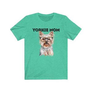 May include: A teal green t-shirt with the text "Yorkie Mom" in black lettering. A watercolor illustration of a Yorkshire Terrier wearing glasses is centered on the shirt.