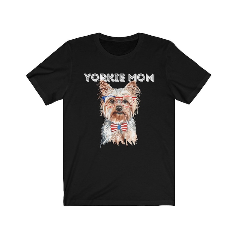May include: Black t-shirt with the text "Yorkie Mom" in white. A watercolor illustration of a Yorkshire Terrier wearing red, white, and blue glasses and a bow tie is centered on the shirt.