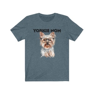 May include: A teal blue t-shirt with the text "Yorkie Mom" in black lettering. A watercolor illustration of a Yorkshire Terrier wearing glasses is centered on the shirt.
