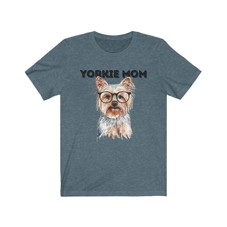 May include: A teal blue t-shirt with the text "Yorkie Mom" in black lettering. A watercolor illustration of a Yorkshire Terrier wearing glasses is centered on the shirt.