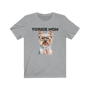 May include: A heather grey t-shirt with the text "Yorkie Mom" printed in black. A watercolor illustration of a Yorkshire Terrier wearing glasses is centered on the shirt.