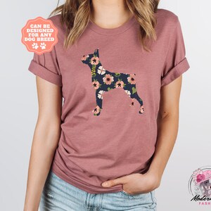 Puede incluir: Una camiseta color malva con una silueta de perro Doberman Pinscher con estampado floral. El texto "Can be designed for any dog breed" est&aacute; en una etiqueta rosa.