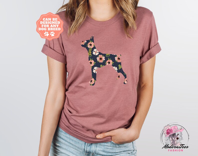 Puede incluir: Una camiseta color malva con una silueta de perro Doberman Pinscher con estampado floral. El texto "Can be designed for any dog breed" est&aacute; en una etiqueta rosa.