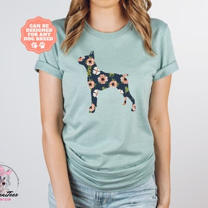 Puede incluir: Una camiseta azul claro con una silueta de perro con estampado floral. El texto "Can be designed for any dog breed" est&aacute; en una pancarta rosa sobre el perro.