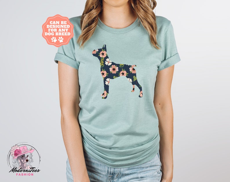 Puede incluir: Una camiseta azul claro con una silueta de perro con estampado floral. El texto "Can be designed for any dog breed" est&aacute; en una pancarta rosa sobre el perro.