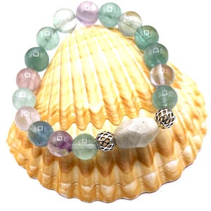 May include: A beaded bracelet featuring a mix of colorful, round gemstones in shades of green, purple, pink, and clear. The bracelet also includes a large, irregular-shaped stone and two silver-colored, decorative beads. The bracelet is displayed on a seashell.