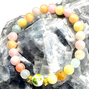 May include: A beaded bracelet featuring a variety of pastel-colored round beads, including pink, yellow, orange, and green. The bracelet also includes a decorative white bead with a floral design and gold accents. The bracelet is displayed on a blue-gray stone surface.