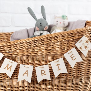 Könnte beinhalten: Ein Weidenkorb mit einem Banner, auf dem "MARIE" in weißen Buchstaben auf hellbraunem Hintergrund steht. Das Banner ist mit Koalabären-Illustrationen verziert. Der Korb ist gefüllt mit Stofftieren, darunter ein grauer Hase und eine weiße Katze.