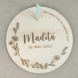 May include: A round wooden plaque with a floral design and the text "Wie sch&ouml;n, dass du geboren bist" in German. The plaque also has the name "Madita" and the date "18. M&auml;rz 2022" in German. A small, light blue candle is in a holder on the plaque.