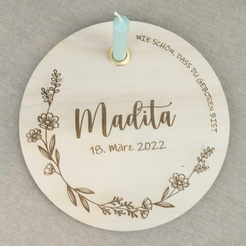 May include: A round wooden plaque with a floral design and the text "Wie sch&ouml;n, dass du geboren bist" in German. The plaque also has the name "Madita" and the date "18. M&auml;rz 2022" in German. A small, light blue candle is in a holder on the plaque.