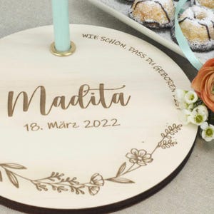 May include: A round wooden plaque with the text "Wie sch&ouml;n, dass du geboren bist" and "Madita 18. M&auml;rz 2022" engraved on it. The plaque has a floral design around the edge and a hole in the center.