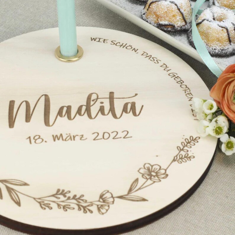 May include: A round wooden plaque with the text "Wie sch&ouml;n, dass du geboren bist" and "Madita 18. M&auml;rz 2022" engraved on it. The plaque has a floral design around the edge and a hole in the center.