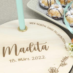 May include: A wooden round plaque with the text "Wie sch&ouml;n, dass du geboren bist" and the name "Madita" with the date "18. M&auml;rz 2022" engraved on it. The plaque has a small hole in the center and a light blue ribbon is threaded through it.