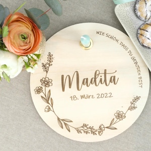 May include: A round wooden plaque with a floral design and the text "Wie sch&ouml;n, dass du geboren bist". The plaque also includes the name "Madita" and the date "18. M&auml;rz 2022".