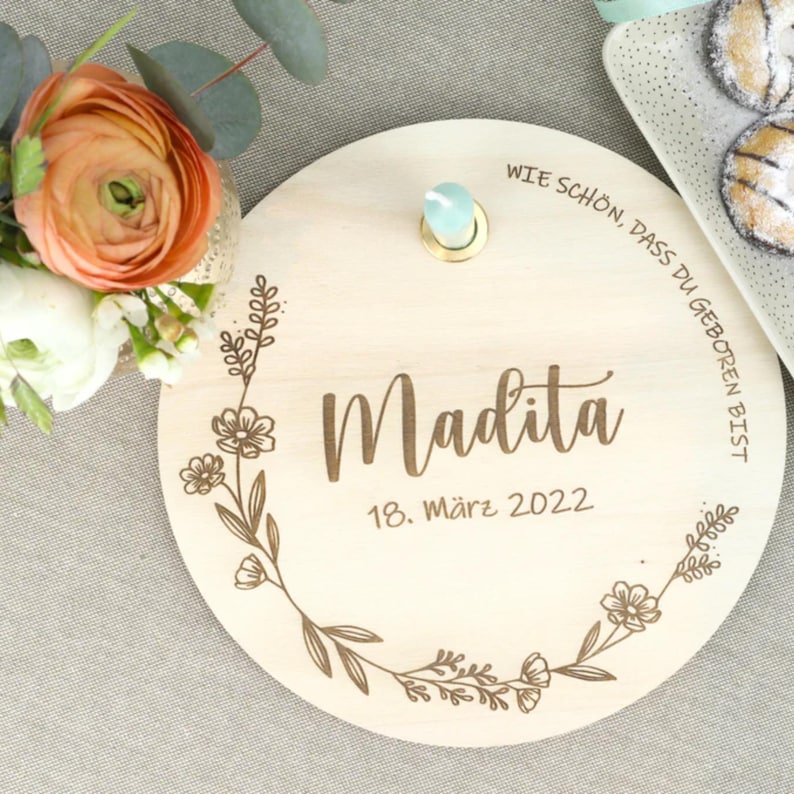 May include: A round wooden plaque with a floral design and the text "Wie sch&ouml;n, dass du geboren bist". The plaque also includes the name "Madita" and the date "18. M&auml;rz 2022".