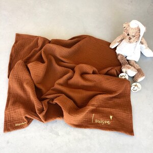 Peut inclure: Une couverture en mousseline bébé de couleur rouille avec le nom "Louison" brodé en fil doré. La couverture est pliée et posée sur une surface grise. Un ours en peluche se trouve en arrière-plan.