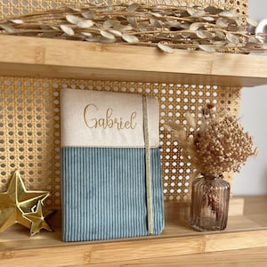 Peut inclure: Un carnet rectangulaire doté d'une couverture bicolore avec une partie supérieure en tissu crème brodée du prénom Gabriel en fil doré et une partie inférieure en velours côtelé bleu, maintenu par un élastique vertical doré.