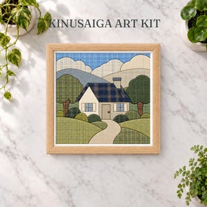 May include: A square Kinusaiga art kit featuring a fabric-pieced landscape of a house with a blue plaid roof, a chimney, and a winding path. The scene includes green hills, trees, and blue mountains under a sky, all encased in a light wood frame.