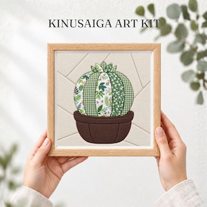 Può includere: Un kit d'arte Kinusaiga con una cornice quadrata in legno contenente un progetto artigianale in tessuto. L'opera raffigura un cactus verde rotondo in un vaso marrone, costruito con vari segmenti di tessuto a motivi su uno sfondo color crema.