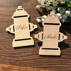 May include: Two light-colored wooden cutouts shaped like fire hydrants. Each cutout features a horizontal slot at the top and bottom, with the names Sofia and Nicole engraved in a cursive script on the center of each respective wooden piece.