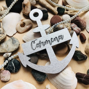 Peut inclure: Décoration argentée en forme d'ancre avec le nom "Carmena" gravé. L'ancre est entourée de coquillages, de perles et de petites pierres sur une surface en bois clair. Décoration sur le thème nautique.