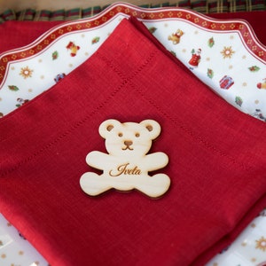 Könnte beinhalten: Eine rote Serviette mit einem hölzernen Teddybär-Platzkarte, auf der "Iveta" steht. Die Serviette liegt auf einem weißen Teller mit rotem Muster.
