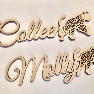 Op de afbeelding: Twee houten naamuitsparingen, "Colleen" en "Molly", in een cursief lettertype. Elke naam gaat vergezeld van een houten luipaardsilhouet met zwarte vlekken. De items zijn gemaakt van lichtgekleurd hout.