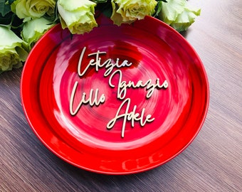 May include: A red plate with four wooden place cards with the names Letizia, Ignazio, Lillo, and Adele written on them.