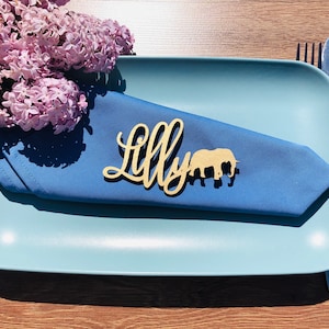 May include: A wooden place card with the name "Lilly" written in cursive script. An elephant silhouette is next to the name. The place card is on a blue napkin on a light blue plate.