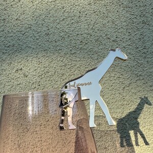 Può includere: Un ciondolo per bevande a forma di giraffa argentata con il nome "Julianne" inciso, appoggiato sul bordo di un bicchiere. Il ciondolo proietta un'ombra su un muro strutturato. Il ciondolo è un oggetto decorativo.