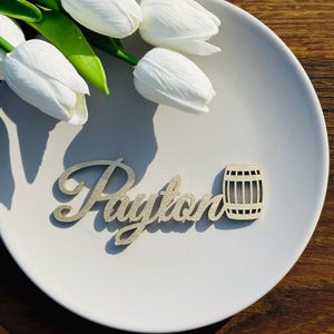 May include: Wooden name card with the name "Payton" and a small wooden barrel design. The card is on a white plate with white tulips in the background.