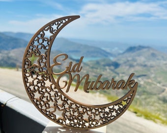 May include: A wooden crescent moon cutout with stars and the words "Eid Mubarak" in a decorative font. The moon is sitting on a wooden stand against a backdrop of a mountain range and a blue sky with white clouds.