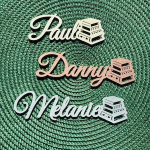 Può includere: Tre ritagli di nomi in legno: "Paul" in crema, "Danny" in oro rosa e "Melanie" in glitter argento. Ogni nome è abbinato a una pila di libri. Lo sfondo è una superficie verde strutturata.
