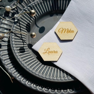 May include: Two hexagonal wooden place cards with the names "Mika" and "Laura" written in gold lettering. The place cards are on a white napkin and a gray patterned plate.