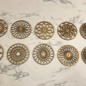 May include: Ten wooden coasters with intricate laser-cut designs. Each coaster features a different geometric pattern, including spirals, stars, and sunbursts. The coasters are all made of natural wood and have a light brown color.